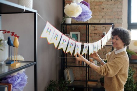 Foto de Mujer joven decorando la habitación para la fiesta de cumpleaños colgando cartel de feliz cumpleaños entre dos estantes - Imagen libre de derechos