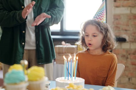 Foto de Niño pequeño con el pelo largo en sombrero de fiesta sentado en la mesa con pastel de cumpleaños y soplando velas en él - Imagen libre de derechos