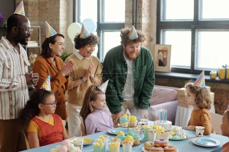 Foto de Grupo de niños y adultos en sombreros de fiesta aplaudiendo mientras niño soplando velas en pastel de cumpleaños - Imagen libre de derechos
