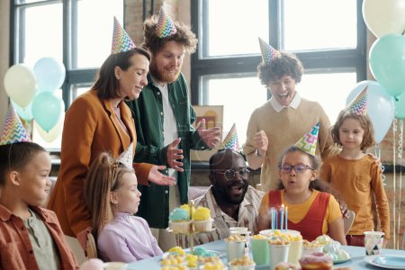 Téléchargez les photos : Petite fille en chapeau de fête soufflant des bougies sur le gâteau d'anniversaire avec ses parents et amis la félicitant avec anniversaire - en image libre de droit