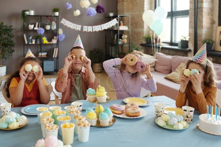 Group of children holding desserts like eyes on their faces and posing at camera while sitting at table with dessert during birthday party
