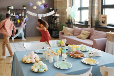 Foto de Imagen de mesa con postres y comida dulce con grupo de niños corriendo y divirtiéndose juntos en segundo plano en la fiesta - Imagen libre de derechos