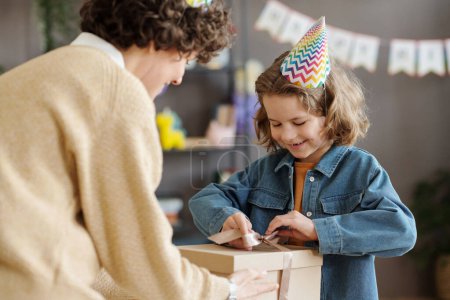 Foto de Feliz niño en el sombrero de fiesta apertura de la caja de regalo dando por su madre en el cumpleaños - Imagen libre de derechos