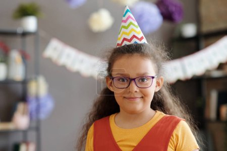 Photo for Portrait of little girl in eyeglasses and party hat smiling at camera at birthday party - Royalty Free Image