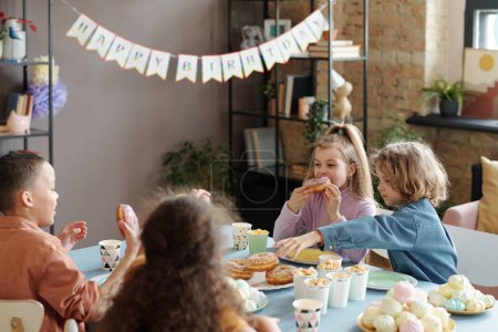 Téléchargez les photos : Groupe d'enfants assis à table et dégustant des beignets et autres aliments sucrés pendant la fête d'anniversaire à la maison - en image libre de droit