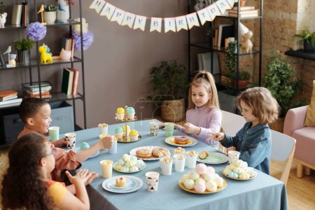 Photo for Group of children playing rock paper scissors game while sitting at table with sweet food in living room at birthday party - Royalty Free Image