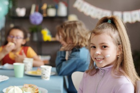 Téléchargez les photos : Portrait de petite fille avec de la crème sur le visage souriant à la caméra alors qu'elle était assise à table avec ses amis et mangeant du dessert à la fête - en image libre de droit