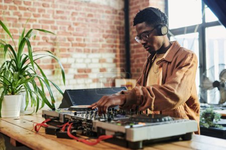 Téléchargez les photos : Jeune homme noir sérieux avec des écouteurs sur la tête mélangeant les sons tout en restant près des platines tournantes et en faisant de la musique en studio - en image libre de droit