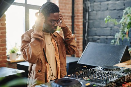 Foto de Joven hombre negro poniendo auriculares en su cabeza mientras está de pie junto a la mesa con equipo musical en la sala de estar del apartamento loft - Imagen libre de derechos