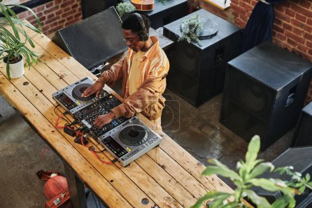 Téléchargez les photos : Vue d'ensemble du jeune DJ masculin afro-américain debout près d'une longue table en bois avec dj set et la création de nouvelles musiques - en image libre de droit