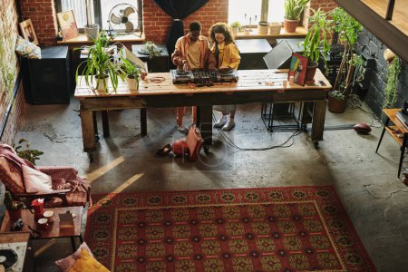 Foto de Joven pareja interracial en ropa casual de pie junto a una larga mesa de madera con cabina de dj y mezclando sonidos mientras crea nueva música - Imagen libre de derechos