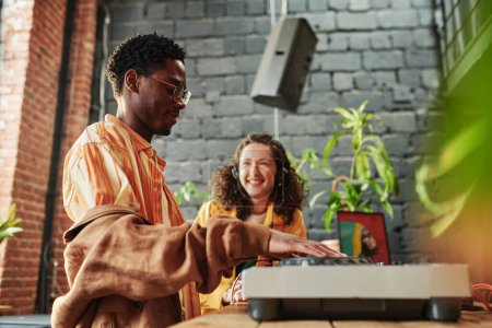 Photo for Happy black man adjusting dj controller while creating new music against his cheerful girlfriend while spending time with her at home - Royalty Free Image
