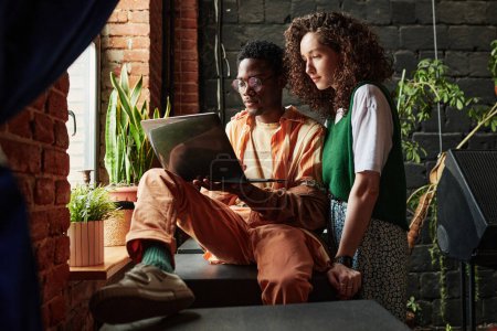 Téléchargez les photos : Jeune couple interracial travaillant dans le filet tandis que l'homme tenant un ordinateur portable et sa petite amie debout à côté de lui devant la fenêtre en studio - en image libre de droit