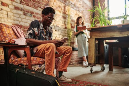 Photo for Young serious black man in casualwear using mobile phone while sitting in armchair against his girlfriend reading book while standing by wall - Royalty Free Image