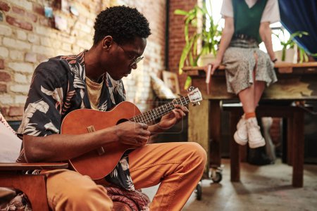 Photo for Young man in casual shirt and pants playing guitar while sitting in armchair in front of his girlfriend wearing skirt and waistcoat - Royalty Free Image