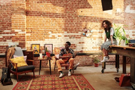 Photo for Young female in casualwear sitting on table and listening to her African American boyfriend playing guitar and singing - Royalty Free Image