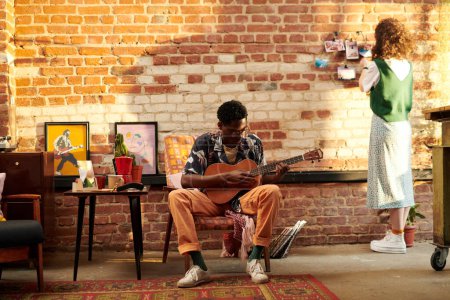 Téléchargez les photos : Jeune homme assis dans un fauteuil et jouant de la guitare acoustique tandis que sa petite amie debout devant un mur de briques et regardant des photos - en image libre de droit