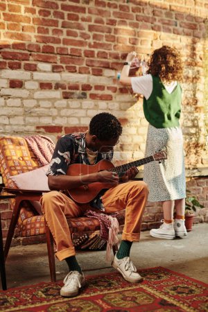 Foto de Joven tocando la guitarra acústica contra su novia de pie frente a la pared de ladrillo en el estudio o sala de estar del apartamento loft moderno - Imagen libre de derechos
