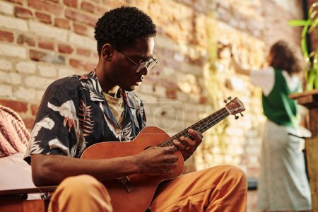 Young black man with acoustic guitar sitting in armchair against his girlfriend standing in front of brick wall in living room of loft apartment