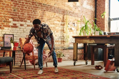 Téléchargez les photos : Jeune homme afro-américain en tenue décontractée jouant avec la balle sur le tapis dans le salon spacieux de loft appartement avec murs de briques - en image libre de droit