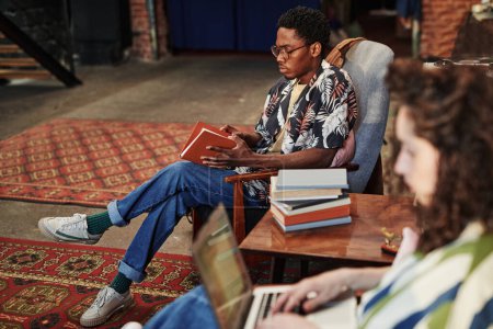 Téléchargez les photos : Sérieux Afro-Américain en chemise élégante et un jean bleu livre d'ouverture en couverture brune tout en étant assis dans un fauteuil dans le salon à loisir - en image libre de droit