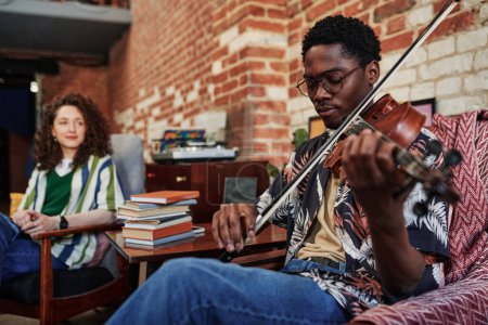 Young man in eyeglases and casualwear sitting in armchair in living room of loft apartment at leisure and playing violin for his girlfriend Poster 645005426