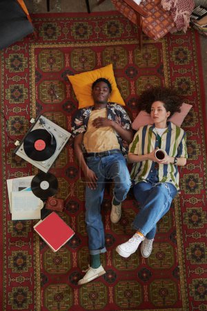 Foto de Joven interracial citas mantener la cabeza en almohadas mientras se relaja en la alfombra roja en el suelo de la sala de estar y escuchar discos vynil - Imagen libre de derechos