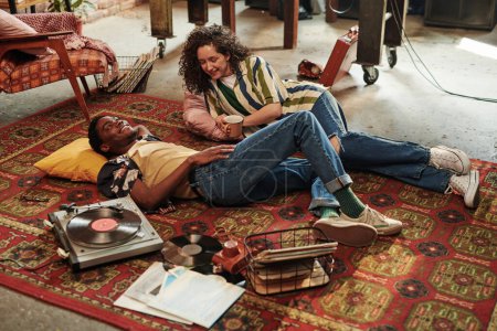 Téléchargez les photos : Joyeux jeune couple en tenue décontractée élégante écoutant des disques vynil et parlant tout en se relaxant sur le tapis rouge sur le sol du salon - en image libre de droit