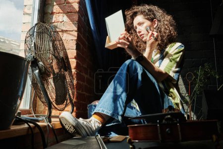 Foto de Mujer joven en jeans azules y zapatos de goma blancos aplicando rubor en sus mejillas frente a la ventana por la mañana mientras se mira en el espejo - Imagen libre de derechos