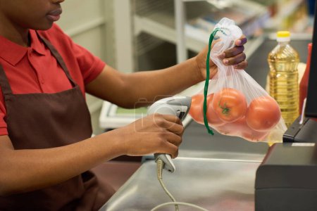 Téléchargez les photos : Jeune femme noire en chemise rouge et tablier marron scannant les tomates fraîches dans un sac de cellophane au comptoir de caisse dans un supermarché - en image libre de droit