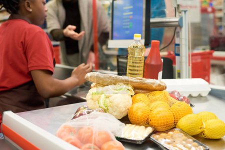 Téléchargez les photos : Légumes frais emballés, fruits et autres produits alimentaires scannés par une caissière au service de l'un des acheteurs dans un supermarché - en image libre de droit