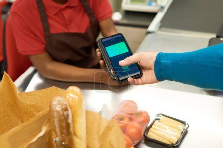 Téléchargez les photos : Jeune afro-américain détenant terminal de paiement lors du paiement sans contact de produits alimentaires par le consommateur avec smartphone - en image libre de droit