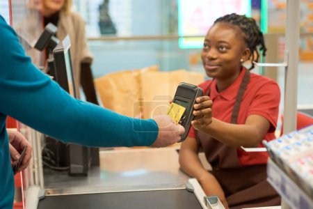 Foto de Comprador moderno que paga por las mercancías elegidas con tarjeta de crédito mientras lo sostiene por terminal de pago en poder de la joven vendedora afroamericana - Imagen libre de derechos