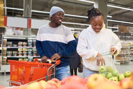 Téléchargez les photos : Heureux jeune couple afro-américain de zoomers choisir des fruits frais tout en restant debout par affichage dans le département d'épicerie du supermarché - en image libre de droit