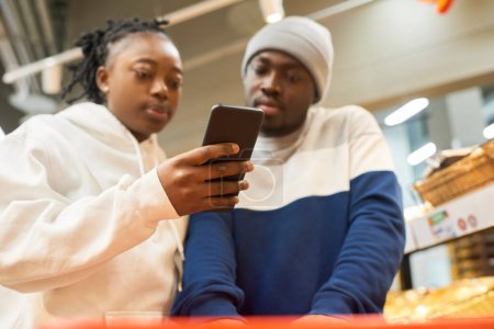 Foto de Joven mujer negra con smartphone mostrando la lista de compras a su novio mientras elige productos alimenticios en el supermercado - Imagen libre de derechos