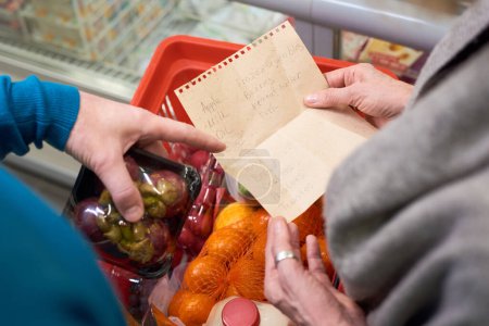 Photo for Overview of hand of mature man pointing at shopping list held by his wife over cart with fresh packed fruits and other food products - Royalty Free Image