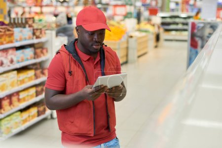 Téléchargez les photos : Jeune travailleur masculin du supermarché regardant l'écran de la tablette numérique tout en vérifiant les prix des produits alimentaires dans le département de l'épicerie - en image libre de droit