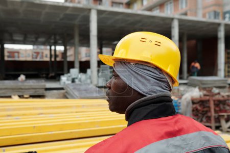 Téléchargez les photos : Homme noir mature en casque de protection et vêtements de travail debout sur le chantier de construction en face de la structure inachevée du bloc d'appartements - en image libre de droit