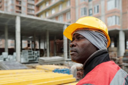 Photo for African American male builder in workwear and hardhat looking aside while standing against unfinished building at construction site - Royalty Free Image