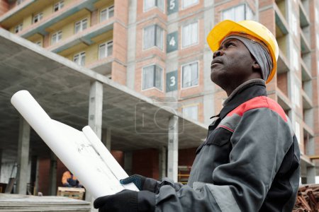 Foto de Ingeniero afroamericano en ropa de trabajo mirando hacia arriba en el condominio de gran altura mientras está parado en el área de construcción contra la construcción - Imagen libre de derechos