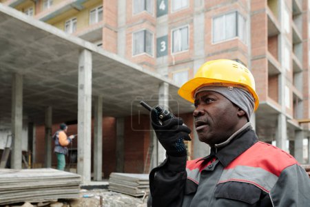Téléchargez les photos : Constructeur âgé en casque rigide et vêtements de travail parlant en talkie-walkie tout en travaillant sur le chantier de construction contre la structure en béton inachevé - en image libre de droit
