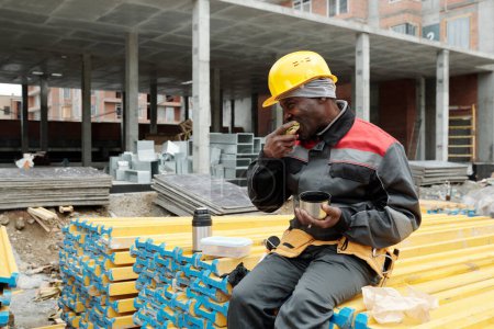 Téléchargez les photos : Homme noir mature affamé en hardhat et vêtements de travail manger un sandwich et boire du thé chaud tout en étant assis sur une pile de matériaux de construction - en image libre de droit