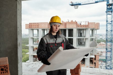 Photo for Young female worker of construction site with blueprint standing inside unfinished building against crane and another structure - Royalty Free Image