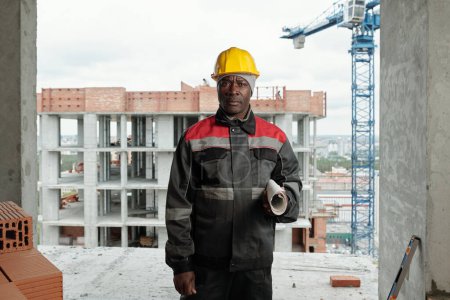 Téléchargez les photos : Constructeur afro-américain mature avec un plan laminé regardant la caméra tout en se tenant contre des bâtiments inachevés sur le chantier de construction - en image libre de droit