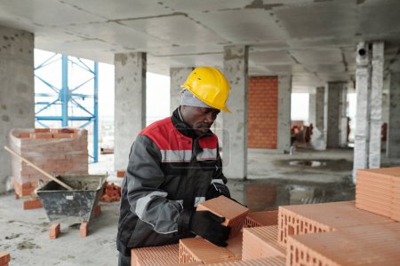 Téléchargez les photos : Mature homme noir en vêtements de travail et casque de protection mettre des briques en pile tout en travaillant dans un bâtiment inachevé sur le chantier de construction - en image libre de droit