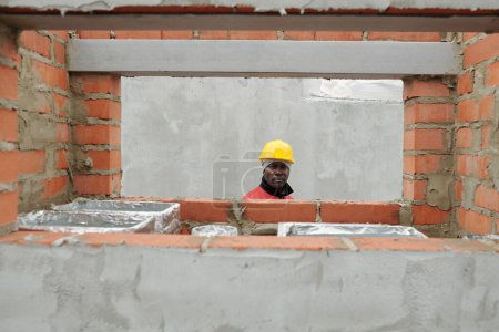 Foto de Constructor afroamericano maduro en casco protector y ropa de trabajo de pie por pared de ladrillo con marco de ventana y materiales de construcción - Imagen libre de derechos