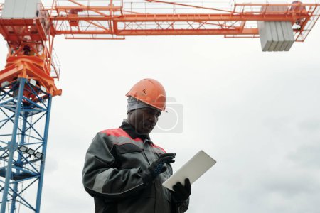 Téléchargez les photos : Homme noir mature en gants, vêtements de travail et casque feuilleté défilant dans l'information en ligne tout en utilisant une tablette sur le chantier de construction - en image libre de droit