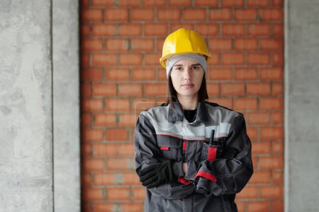 Photo for Young serious female engineer in protective helmet and workwear looking at you while standing in front of camera against brick wall - Royalty Free Image