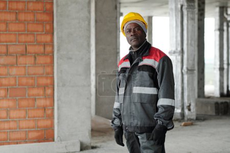 Téléchargez les photos : Constructeur afro-américain âgé ou contremaître en uniforme et casque debout contre des colonnes de béton à l'intérieur du bâtiment inachevé - en image libre de droit