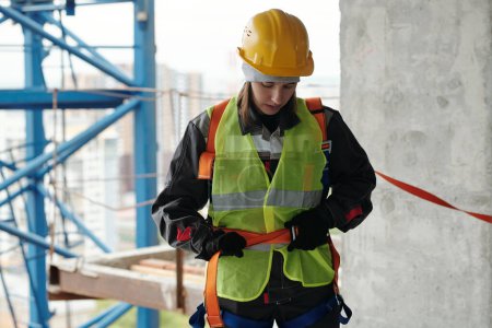 Photo for Young female builder in protective helmet and reflective vest fastening safety belt on waist before building work at construction site - Royalty Free Image